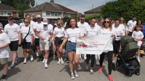 A group of people in white t-shirts take part in a walk, starting in a car park with houses visible behind them.