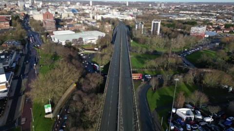 An aerial photograph of the Gateshead flyover. The bridge on the A167 runs towards Gateshead town centre and the Tyne Bridge. A car dealership can be seen to the right of the bridge while commercial buildings stand to the left. Newcastle can be seen in the distance.