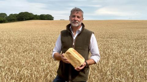 A photo of a man standing in a field with a loaf of bread, he is looking directly down into the camera. He is wearing a shirt and a vest jacket.