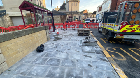 A newly-laid footpath with a works van in the road and red barrier gates surrounding the area, with the town in the background