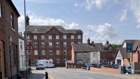 A road in the centre of a village curves around to the left. Buildings are either side and cars can be seen driving along the road or parked in a car park on the right.