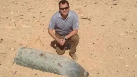 Bomb disposal expert Ben Remfrey wearing sunglasses, a grey polo shirt, brown trousers and brown shoes while kneeling next to a large unexploded grey-coloured bomb in a sandy area.