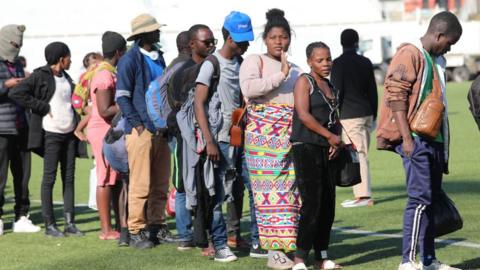 People standing in a queue on some grass. Some are carrying backs on their backs and wearing hats.