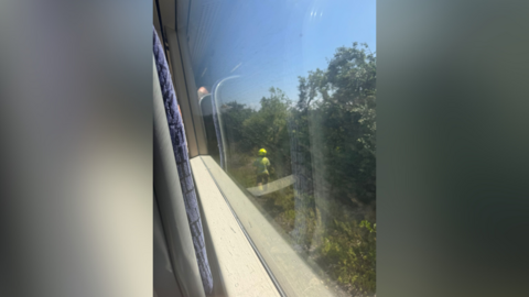 A photo looking out of the window of a train. A man in yellow helmet is stood next to the tracks.