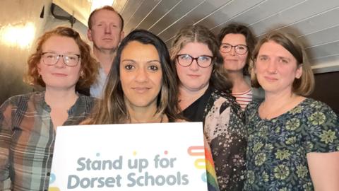 A group of parents - five women and a man - pose for the camera with a board bearing the logo of their campaign group: Stand up for Dorset Schools
