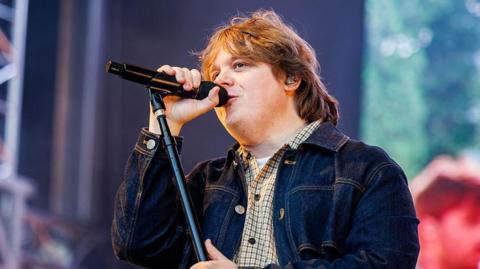 Lewis Capaldi singing onstage - he has light brown hair and a denim jacket over a chequed shirt. 