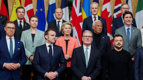 Sir Keir Starmer, Emmanuel Macron and Volodymyr Zelensky surrounded by other world leaders in a group photo in front of their nation's flags at Lancaster House.