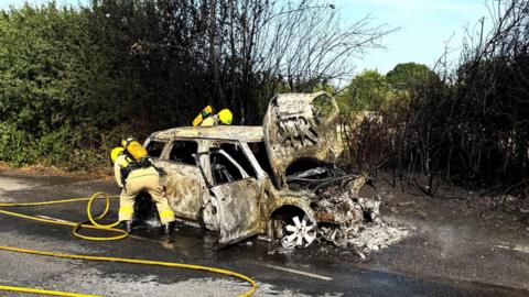 A once white mini countryman car sits by the side of a road charred and blackened by fire. Its bonnet is up and the front of the vehicle has melted. The wheels have also blown out and melted. Behind it is charred and blackened hedges . Two firefighters have yellow hoses and are spraying the car