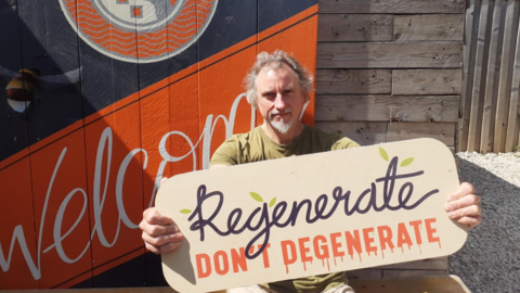 A man with wayward mid-length wavy grey hair holds up a sign that reads "Regenerate don't regenerate" as he sits outside on a gravelled area in the sunshine. The word "Welcome" is painted on a shed behind him.