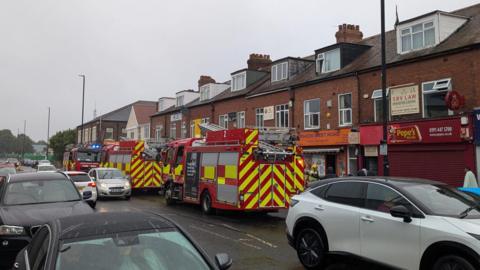 Three fire engines parked outside Krakow West Road shop in Newcastle. A firefighter is standing outside the building. Traffic is moving slowly past the three engines which are parked in one lane of the road.