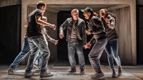 Image shows a group of people all grouped together, all facing into a circle. They are on stage and the light falls on their faces so you can see their features clearly. They are all wearing casual clothes - jackets, jeans. 