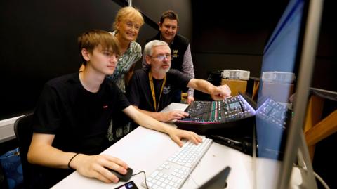 Four people working together at computers and looking at something on a screen. A man is pointing something out to the other three, who include a younger man, a woman and another man. 