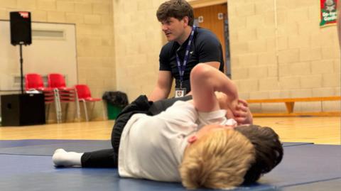 Two boys in white and black sports kit are wrestling on blue mats in the foreground. Behind them sitting on the mat is coach Callum Perry wearing a black t-shirt. They are in a school sports hall.