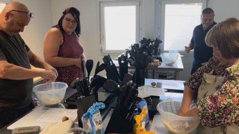 A group of people in a kitchen. The tables have cooking implements on them as well as mixing bowls which the people are stood over making socnes in them.