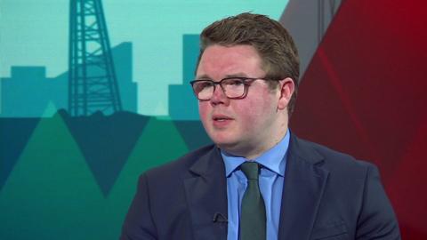 Councillor Antony Mullen sits in a TV studio wearing a blue suit and green tie. He has thick wavy brown hair and wears black-rimmed glasses. The teal and red backdrop of the BBC Politics North studio can be seen behind him. 