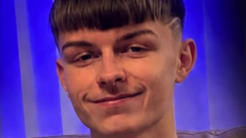 A teenage boy smiling at the camera. He has short brown hair and is against a purple background.