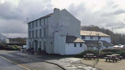 Three-storey old white building with the name Yew Tree emblazoned on the side. Wooden benches and tables surround it with a road in front.