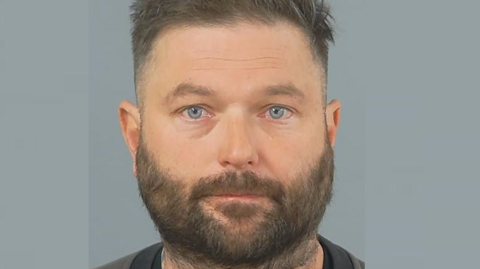 Darren Silvester has a blank expression in a police photo. He has brown hair, closely shaved at the sides, and a beard.