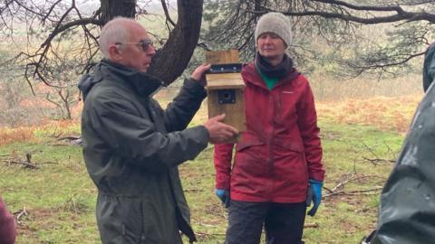Two bird enthusiasts holding aloft a bird box - the man is wearing a dark coat and the woman a red coat with a beanie hat and gloves 