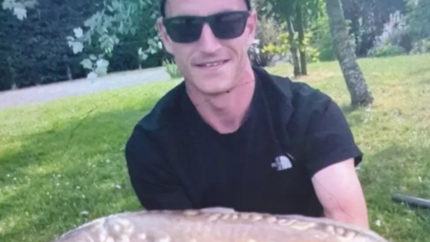 Louis Price, wearing a black t-shirt and dark sunglasses holding a fish in a grassy area