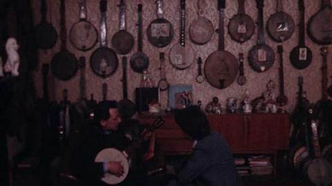 Reuben and the reporter are visible at the bottom of the image. Reuben is holding a banjo.  Behind there are many banjos displayed on the wall.