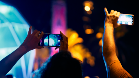 Two people in a darkly lit nightclub holding their phones in landscape towards the stage.
