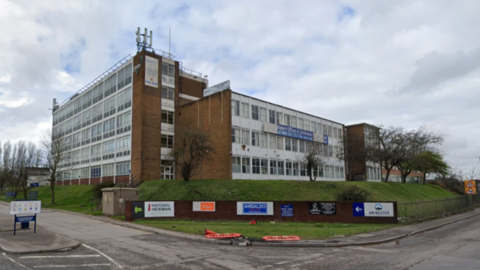 The large building stands on slightly raised ground on a corner with business names on signs below on a grass verge. The building straddles the corner plot.