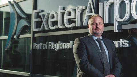 Stephen Wiltshire, Managing Director of Exeter Airport, stands in front of a sign which reads Exeter Airport and appears to be on the side of a building. His expression is serious as he stands to the right looking at the camera.