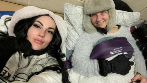Elaine wearing a white hoodie, taking a selfie with her mum Linda in a grey jumper and a beige hat.
