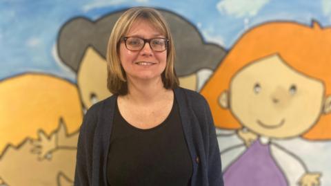 A woman wearing a black top and navy blue cardigan stands in front of a painted wall. She wears glasses and has shoulder length dark-blonde hair
