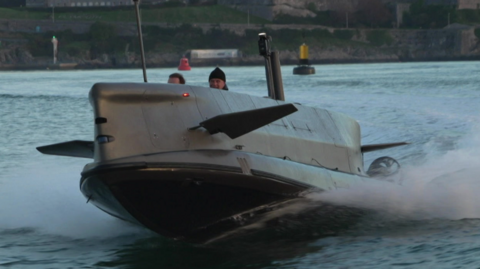 The image shows a sleek, military-grade tactical diving vehicle speeding across the water. Its design is streamlined for high performance, with a dark hull and angular upper structure. The craft is partially surfaced, creating a spray as it moves at high speed, and features antennas and periscopic elements on top. In the background, there is a coastline with historic stone buildings and green areas, along with navigation buoys marking the waterway.