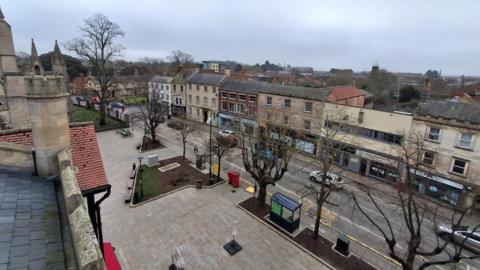 Arial view of Sleaford town centre. There is a row of three-storey buildings, with businesses occupying the lower floors. Cars are also visible on the road, with trees and a bus shelter in the foreground.