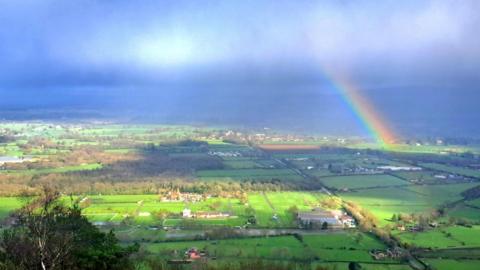 Blue skies with wispy cloud over the Malvern countryside. Some sunshine is peaking through but a vivid rainbow can also be seen on the left. 