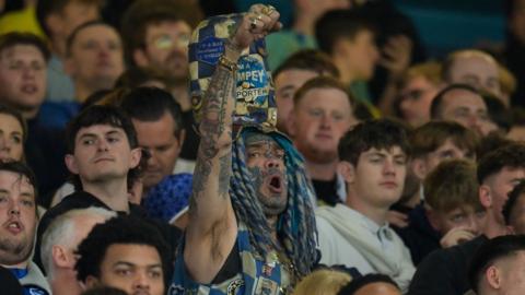 A Portsmouth fan wears white and blue dreadlocks and huge hat with Pompey badges. His face is tattooed and he raises his fist in the air chanting.