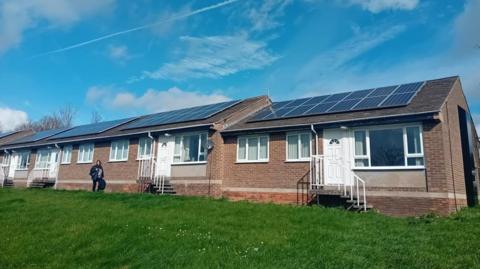 The image shows a row of single‑storey brick homes, each with white doors and windows, set on a grassy slope. The rooftops are fitted with large solar panel arrays stretching across the length of the buildings. The sky above is bright and blue with scattered clouds. A person is walking along the grass in front of the homes.