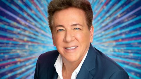 Ross King is smiling at the camera. He has short, dark hair and is wearing a white shirt and blue suit jacket. The background is different shades of blue and red lines with gold sparkles.