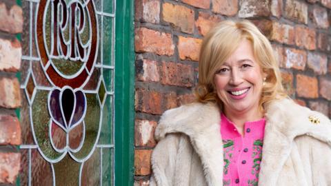 Katy Cavanagh as Julie Carp in Coronation Street. She is wearing a white furry jacket and a pink top.