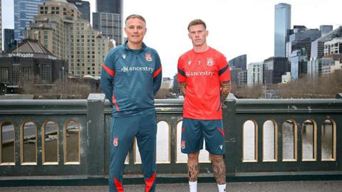 Phil Parkinson stood in a petrol-coloured Wrexham tracksuit with arms behind back next to James McClean in shorts and red training top. They are stood on a bridge with the Melbourne cityscape behind them. 