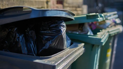 A close-up picture of three bins - one black bin and two green bins - which are both overflowing with rubbish or black bin bags.