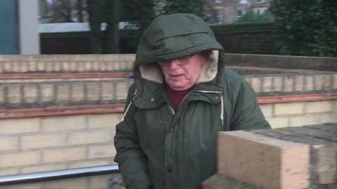 Dennis Finbow is wearing a green coat with the hood up covering his face. His head is angled towards the floor and he looks to be walking. He also wears glasses and a red jumper. 