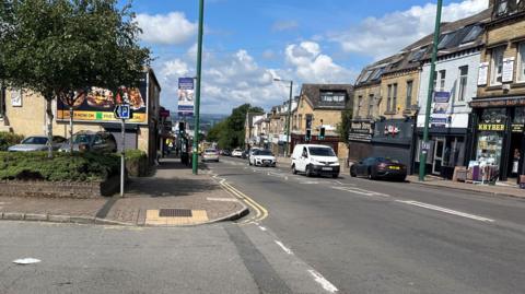 An image of Leeds Road in Bradford, with a car park on the left side and traffic using both lanes on the right side of the photo. 