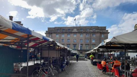 The Market Square on a cloudy day with people sat at some of the outdoor stalls. They have different coloured tarp roofs and are in the shadow or the large Guildhall building. It is light bricked and towers over the square. The market appears shit - apart from a few food stalls.