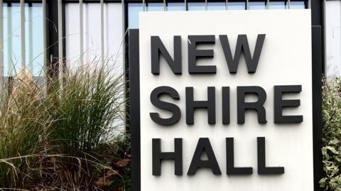 Sign saying "New Shire Hall" in black capital letters outside a modern glass building, surrounded by plants and shrubs.