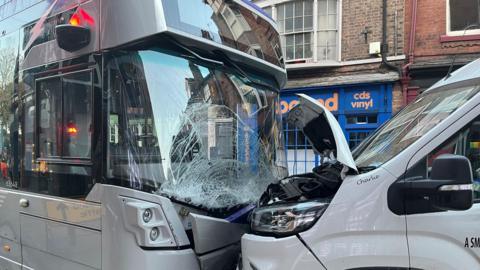 A white single decker bus crashed into the front of a white van. The bus has a cracked windscreen and the van has damage to its bonnet.
