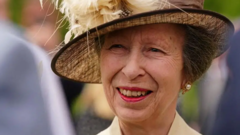 A close-up of Princess Anne smiling. She had short brown hair which is tucked under a brown hat.