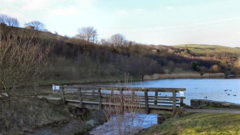 The bridge over the River Medlock at the southern end of the Strinesdale Lower Reservoir. There are hills in the background and a wooden bridge in the foreground.