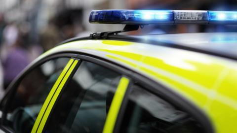 A stock image of an English police car, with blue and yellow livery and a flashing blue light on the roof