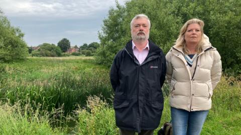 Two people stand side‑by‑side in outdoor jackets beside a grassy, pond‑like area with tall reeds, trees, and distant houses under a cloudy sky.