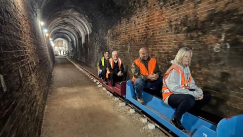 A miniature railway with four people wearing fluorescent jackets in a long tunnel