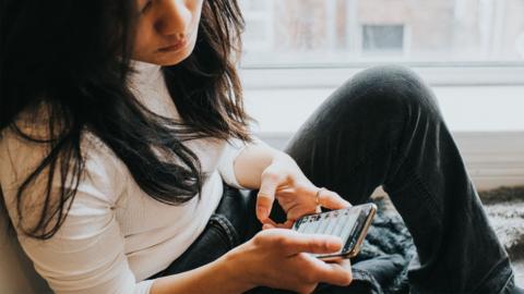 A woman with long black hair wearing a white top looks down at a phone in her hands.